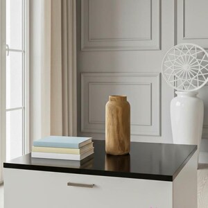 May include: A modern living room scene featuring a white and black cabinet with a glossy black top. A stack of books and a wooden vase sit on the cabinet. A white vase with a decorative top and a window with beige curtains are in the background.