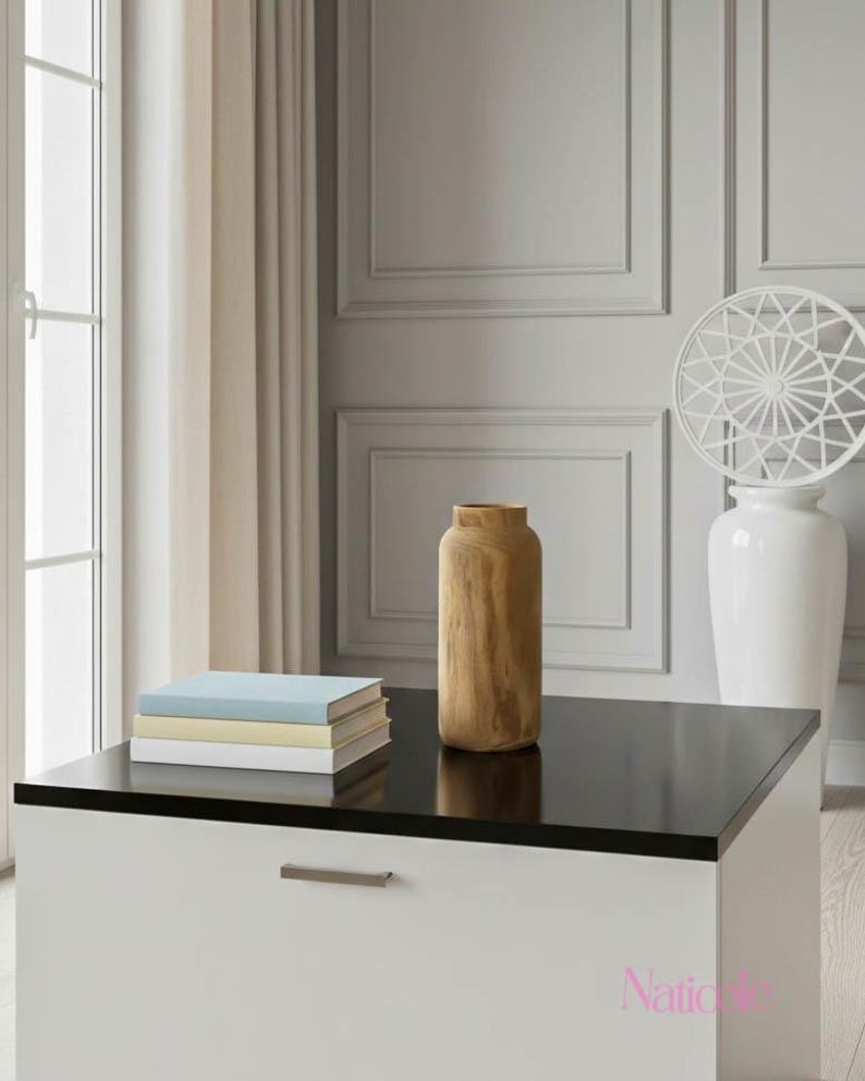 May include: A modern living room scene featuring a white and black cabinet with a glossy black top. A stack of books and a wooden vase sit on the cabinet. A white vase with a decorative top and a window with beige curtains are in the background.