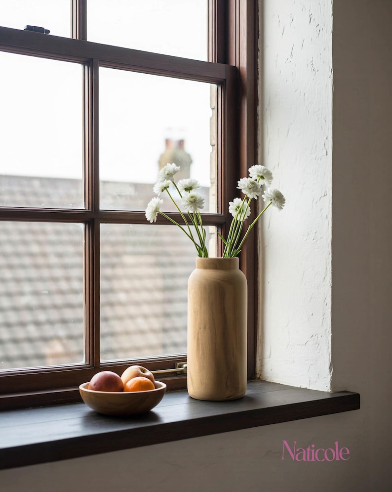 May include: A wooden vase holding white flowers sits on a dark windowsill next to a wooden bowl with fruit. The window frame is dark brown, and the wall is white. The word "Naticole" is in the bottom right corner.