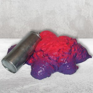 May include: A silver spray paint can lying on its side next to a large blob of pink and purple paint.