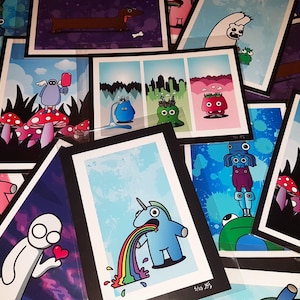 May include: A collection of colourful illustrated prints featuring various characters and designs. The prints are framed in black and white frames and are arranged in a random pattern. Some of the prints feature a unicorn, a creature with a rainbow, and a creature with a crown.