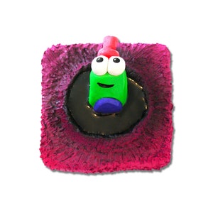 Puede incluir: Un personaje verde y rojo con ojos saltones está sentado en un círculo negro sobre un fondo rosa.