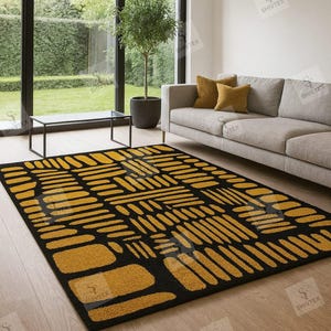 Op de afbeelding: Een zwart en goudkleurig vloerkleed met een geometrisch patroon van langwerpige vormen. Het rechthoekige vloerkleed ligt op een lichte houten vloer in een woonkamer. De kamer heeft een grijze bank en grote ramen.