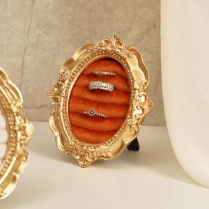 Könnte beinhalten: Ein goldfarbener, verzierter Bilderrahmen mit ovaler Öffnung, der drei Ringe auf einer rostfarbenen Samtoberfläche zeigt. Der Rahmen wird von einem schwarzen Ständer gehalten, der den Schmuck präsentiert.