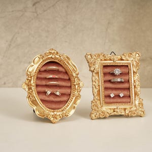 May include: Two ornate gold-colored ring displays. The oval display holds rings and earrings on a pink velvet surface. The rectangular display also holds rings and earrings on a pink velvet surface.