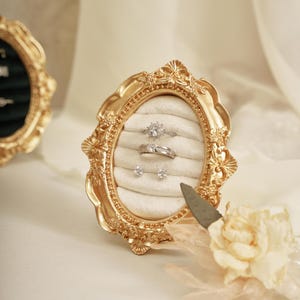 May include: A gold-coloured, ornate picture frame with an oval opening, holding a white velvet ring holder. The holder displays two diamond rings and two diamond stud earrings. A cream-coloured flower and a decorative knife are in the foreground.