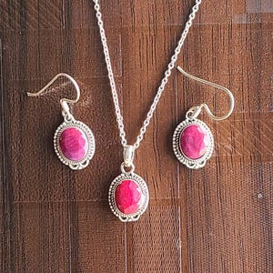 Può includere: Un set di gioielli con una collana d'argento con pendente e orecchini abbinati. Ogni pezzo ha una vivace pietra preziosa ovale rossa incastonata in una cornice d'argento decorativa. Gli orecchini hanno una chiusura a gancio.