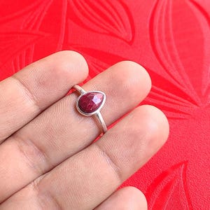 Może przedstawiać: Srebrny pierścionek z fasetowanym, ciemnoczerwonym kamieniem szlachetnym w kształcie łzy. Pierścionek jest umieszczony na czerwonym tle, podkreślając kolor i szlif kamienia. To biżuteria.