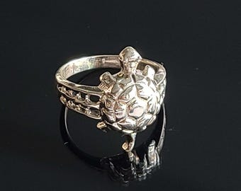 May include: A silver-colored ring featuring a detailed turtle design. The turtle is the focal point, with intricate detailing on its shell and head. The ring band has a decorative pattern. The ring is displayed on a reflective black surface.
