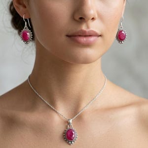 May include: A silver necklace and earrings set with oval, deep pink gemstones. The gemstones are set within a decorative silver border. The earrings are drop style, and the pendant hangs from a delicate chain. The jewellery is displayed against a neutral background.