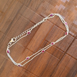 Puede incluir: Una tobillera plateada con piedras preciosas rojas de forma ovalada. La tobillera presenta un diseño de doble cadena con las piedras preciosas espaciadas uniformemente. El cierre es visible, sobre una superficie de madera.
