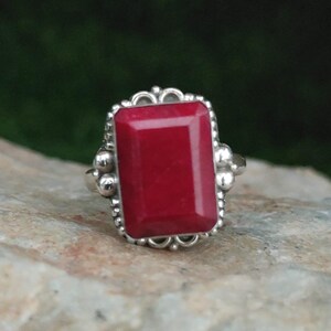 May include: A silver ring featuring a large, rectangular, deep red gemstone set in a decorative metal frame. The setting includes small silver spheres along the sides of the stone, which is polished and smooth with a flat, faceted surface.