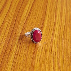 Può includere: Un anello d'argento con una gemma rosso scuro, ovale e sfaccettata. La gemma è incastonata in una lunetta decorativa e smerlata. L'anello è esposto su una superficie di legno con una calda venatura naturale.