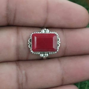 Puede incluir: Un anillo de plata con una piedra roja rectangular facetada, engastada en un marco de metal decorativo con pequeños detalles en forma de cuentas en los bordes. El engaste presenta un diseño calado intrincado alrededor de la piedra.