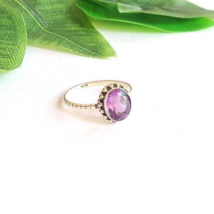 May include: A silver ring featuring an oval, purple gemstone. The ring has a textured band and a decorative bezel. The ring is set against a white background with green leaves.