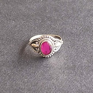 May include: A silver ring featuring a magenta oval gemstone. The ring has an intricate design with small beaded detailing around the gemstone and decorative leaf shapes on the sides. The ring is set against a dark grey background.