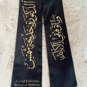 May include: Black graduation sash with gold Arabic calligraphy and the text "Cardiff University School of Medicine 2025". The sash is made of a shiny material, likely for a medical school graduate.