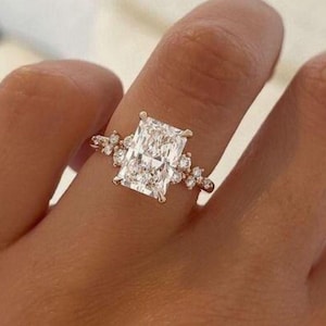 May include: A radiant-cut diamond engagement ring. The ring features a large, rectangular diamond as the centerpiece, set in a delicate band. Smaller accent diamonds are clustered on either side of the main stone, adding sparkle.