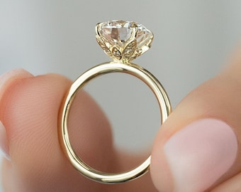Può includere: Un anello di fidanzamento in oro con una grande gemma trasparente. La gemma è incastonata in una montatura decorativa di ispirazione floreale. L'anello è tenuto tra due dita, mettendo in risalto il design e la brillantezza dell'anello.