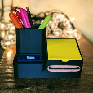 May include: A black desk organizer with compartments for pens, a notepad, and a small drawer. The organizer holds various colored pens and a yellow notepad. A blue USB drive is visible in the drawer. The organizer is on a wooden surface.