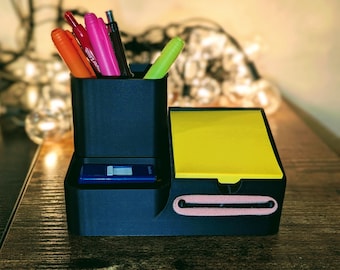 May include: A black desk organizer with compartments for pens, a notepad, and a small drawer. The organizer holds various colored pens and a yellow notepad. A blue USB drive is visible in the drawer. The organizer is on a wooden surface.