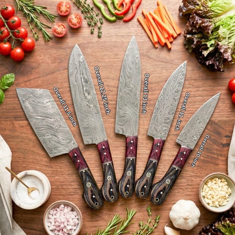 Peut inclure: Un ensemble de cinq couteaux de cuisine en acier damass&eacute; avec des manches en bois sombre, mesurant respectivement 29,2 cm, 31,1 cm, 33 cm, 27,9 cm et 25,4 cm, dispos&eacute;s sur une surface en bois avec des l&eacute;gumes et des herbes frais.