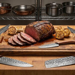 Peut inclure: Un ensemble de quatre couteaux de cuisine avec des lames en m&eacute;tal martel&eacute; et des manches en bois, dispos&eacute;s autour d'une planche &agrave; d&eacute;couper en bois. La planche contient un gros r&ocirc;ti de b&oelig;uf, des tranches de viande et de l'ail r&ocirc;ti.