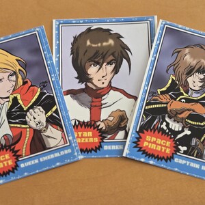 May include: Three trading cards featuring anime characters. The cards have blue borders and include the text "Space Pirate," "Star Blazers," and character names. The characters are depicted in colorful outfits.