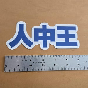 May include: A blue and white sticker with Japanese characters. The sticker is rectangular and features the characters in a bold, sans-serif font. A ruler is placed below the sticker for scale.