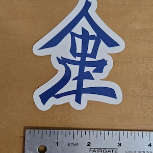 May include: A blue and white sticker featuring a stylized Japanese character. The sticker is on a light brown surface next to a metal ruler with measurements in inches. The ruler reads "FAIRGATE COLD SPRING".