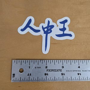 May include: A blue and white sticker with the Chinese characters "人中王" printed on it. A metal ruler is placed below the sticker. The ruler is marked with inches and fractions of an inch.
