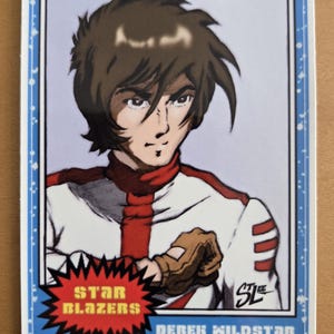 May include: A trading card featuring an anime-style illustration of Derek Wildstar from Star Blazers. He is depicted in a white and red uniform with a determined expression. The card has a blue border and the text "STAR BLAZERS" and "DEREK WILDSTAR".