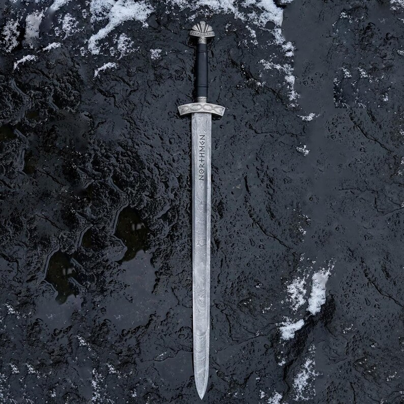May include: A silver sword with a black handle and a decorative hilt, lying on a dark, textured surface with patches of snow. The blade has the word "NORTHMEN" etched vertically.
