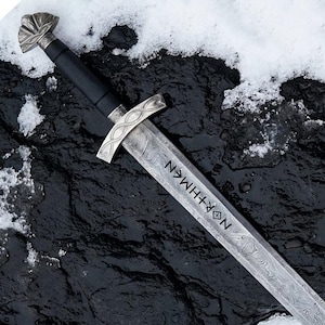 May include: A detailed image of a sword with a patterned blade and a silver crossguard. The handle is black, and the pommel is silver with a decorative design. The blade has the word "NORTHMEN" etched into it. The sword rests on a dark, textured surface with snow.
