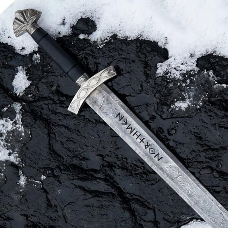 May include: A detailed image of a sword with a patterned blade and a silver crossguard. The handle is black, and the pommel is silver with a decorative design. The blade has the word "NORTHMEN" etched into it. The sword rests on a dark, textured surface with snow.