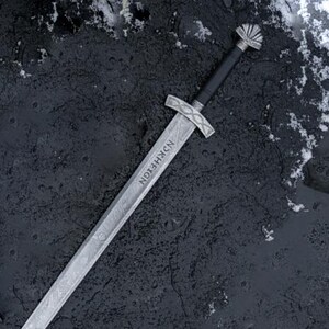 May include: A silver sword with a black handle and a decorative hilt. The blade has a textured pattern and the word "NKRHEION" is etched on the blade. The sword is lying on a dark, textured surface with patches of snow.