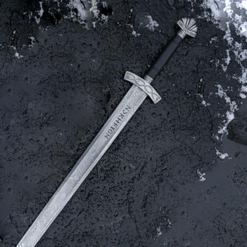 May include: A silver sword with a black handle and a decorative hilt. The blade has a textured pattern and the word "NKRHEION" is etched on the blade. The sword is lying on a dark, textured surface with patches of snow.