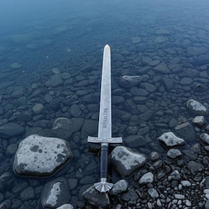 May include: A silver sword with a black handle and a decorative hilt stands upright in shallow water. The blade has etched text. The sword is surrounded by gray rocks in the water.