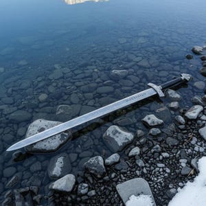 May include: A silver sword with a black handle rests in shallow water among gray rocks. The blade has a patterned design and the word "KOELHIKKEN" is etched on it. The sword's crossguard is silver.