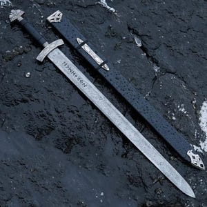 May include: A silver sword with a black handle and a matching sheath. The blade has an etched pattern and the word "NOR+HMAN" is visible. The sheath is black with silver accents.