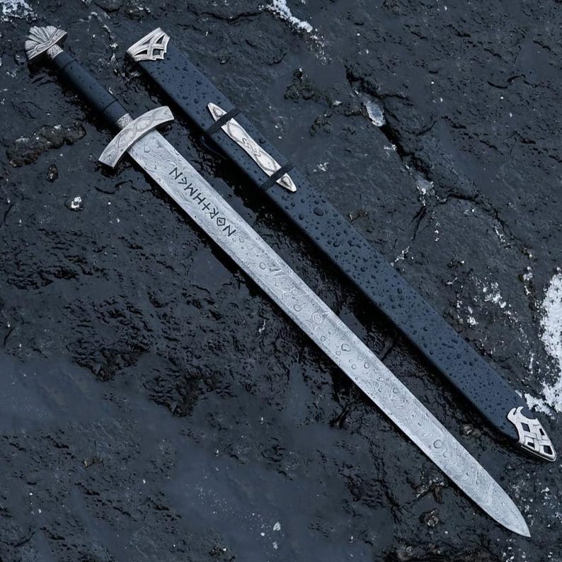 May include: A silver sword with a black handle and a matching sheath. The blade has an etched pattern and the word "NOR+HMAN" is visible. The sheath is black with silver accents.