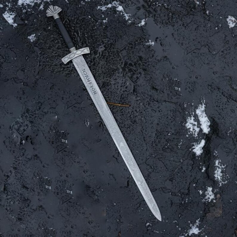 May include: A silver sword with a black handle and a crossguard. The blade has an inscription that reads "NDRKHFYON". The sword is lying on a dark, wet surface with patches of snow.
