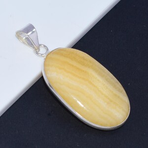 May include: An oval pendant with a polished yellow gemstone, set within a silver-coloured metal frame and bail. The gemstone displays a layered, banded pattern. The pendant is shown against a white and black backdrop.