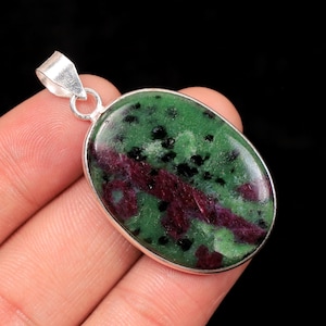 Peut inclure: Pendentif ovale en pierre verte avec des inclusions noires et bordeaux, serti dans un cadre argenté. Le bijou est présenté dans une main sur fond noir, mettant en valeur les motifs et couleurs uniques de la pierre naturelle.