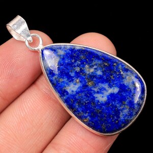 May include: A teardrop-shaped lapis lazuli pendant with a silver-coloured bail. The deep blue stone has flecks of gold and white throughout. The pendant is held against a black background.