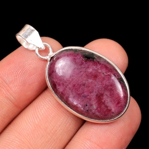 May include: Oval-shaped pendant featuring a polished, deep pink gemstone with black inclusions, set in a silver-toned bezel. The pendant has a loop for a chain and is displayed against a black background.