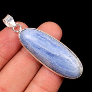 May include: A blue kyanite pendant with a silver-colored bezel and bail. The elongated, oval-shaped stone has a natural, layered appearance with shades of blue and white. The pendant is held against a black background.