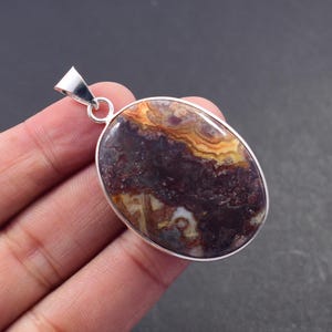 May include: Oval pendant with a polished stone in shades of burgundy, yellow, and cream. The stone is set in a silver-coloured metal frame with a loop for a chain. The pendant is approximately 3.8 cm long.
