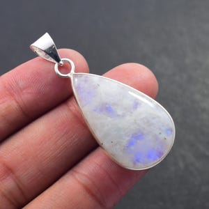 May include: A teardrop-shaped pendant with a polished moonstone and silver bail. The moonstone displays a milky white base with blue and purple flashes. The pendant is held by a hand against a dark background.