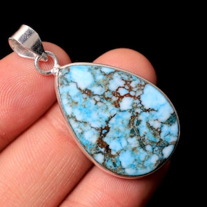 May include: A teardrop-shaped pendant with a silver bail. The pendant features a light blue stone with brown and white veining. The stone is set in a silver bezel. The background is black.